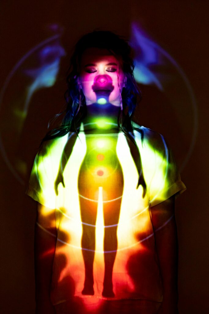 A woman stands in a dark room with a colorful chakra projection on her body.