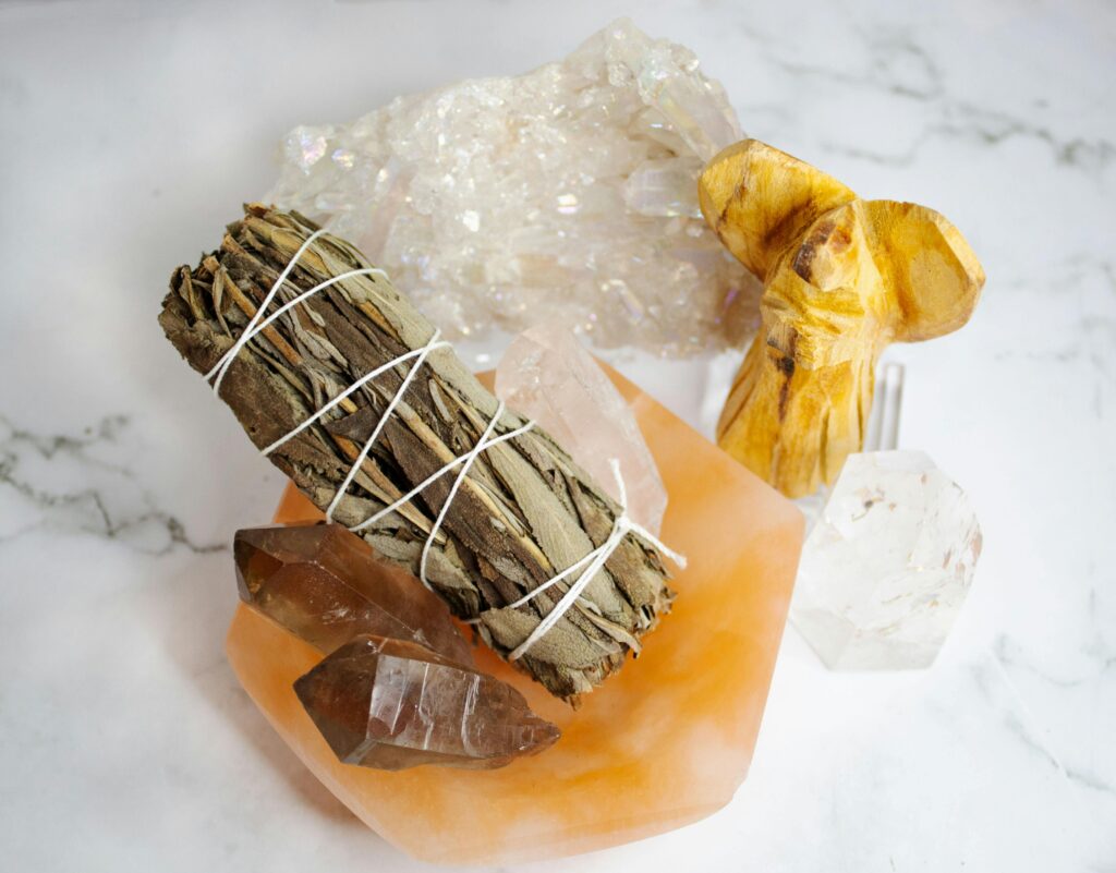A still life of healing crystals, a sage smudge stick, and a wooden angel on a marble surface.